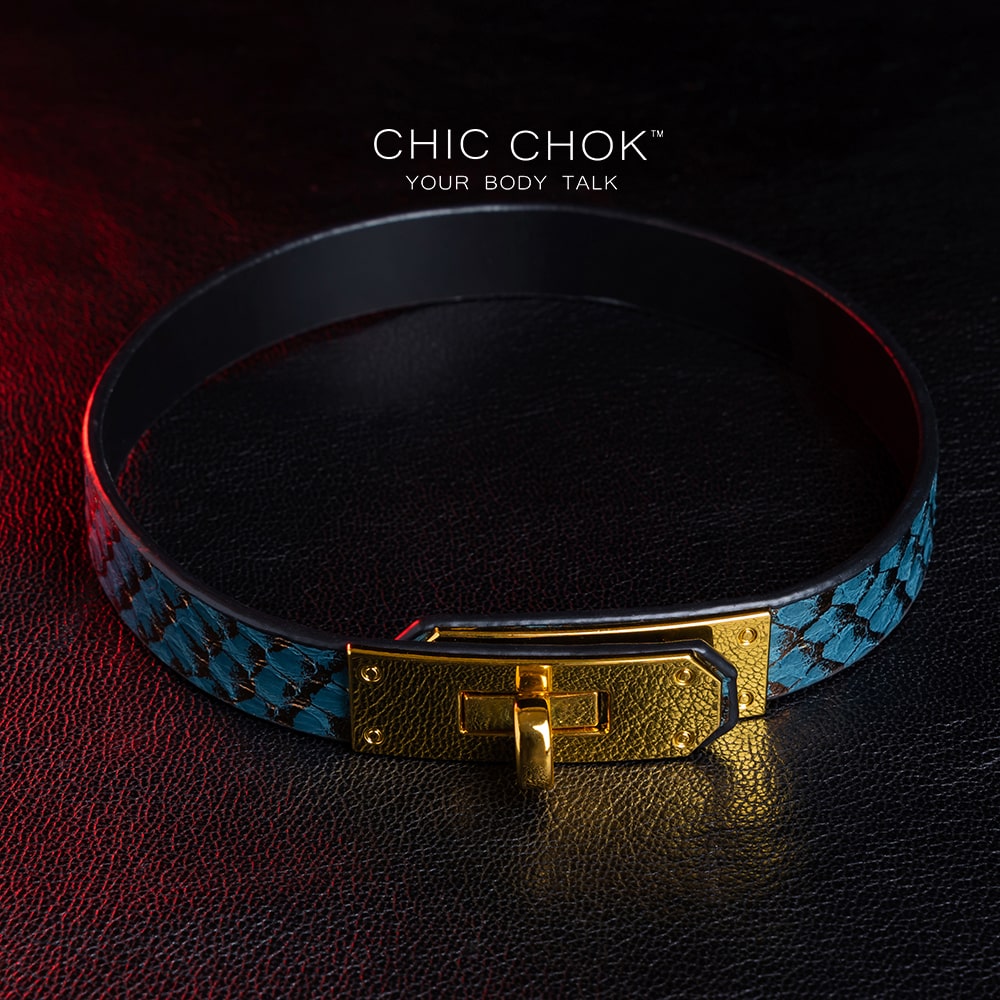 chicchok [Sapphire Moonlight] choker featuring a teal snakeskin-textured band with a gold-plated buckle clasp, displayed on a dark leather surface with dramatic red lighting accents, showcasing its luxurious and edgy design.