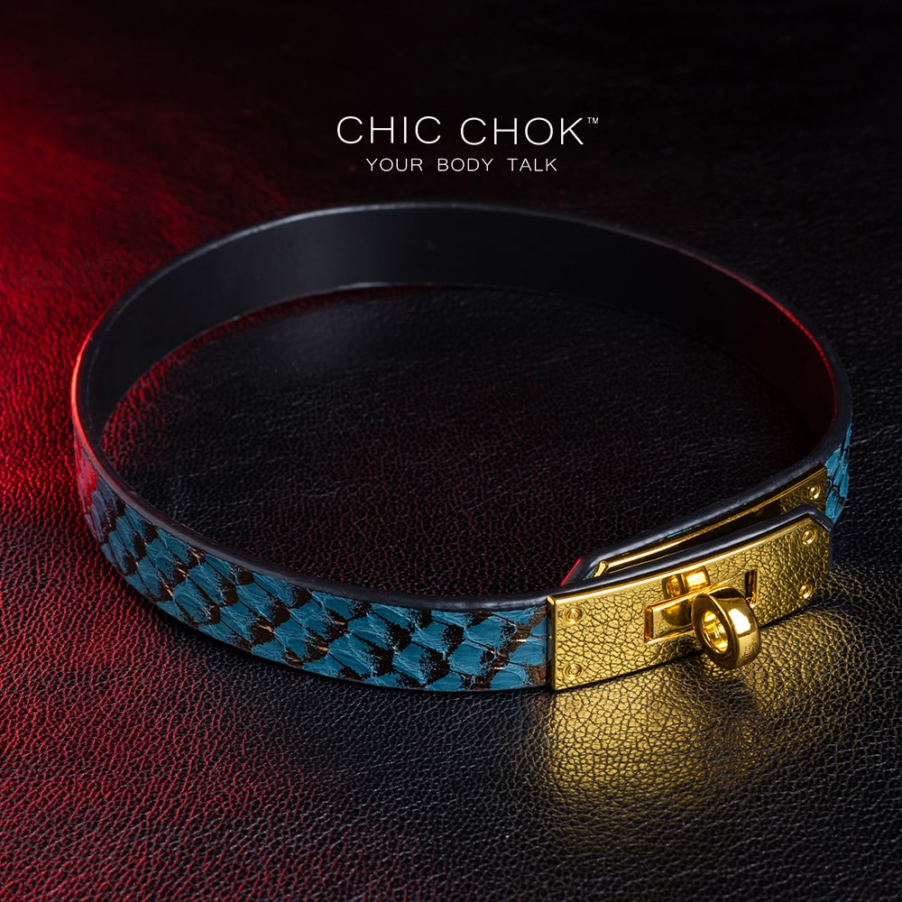 chicchok [Sapphire Moonlight] choker featuring a teal snakeskin-textured band with a gold-plated buckle clasp, displayed on a dark leather surface with dramatic red lighting accents, showcasing its luxurious and edgy design.
