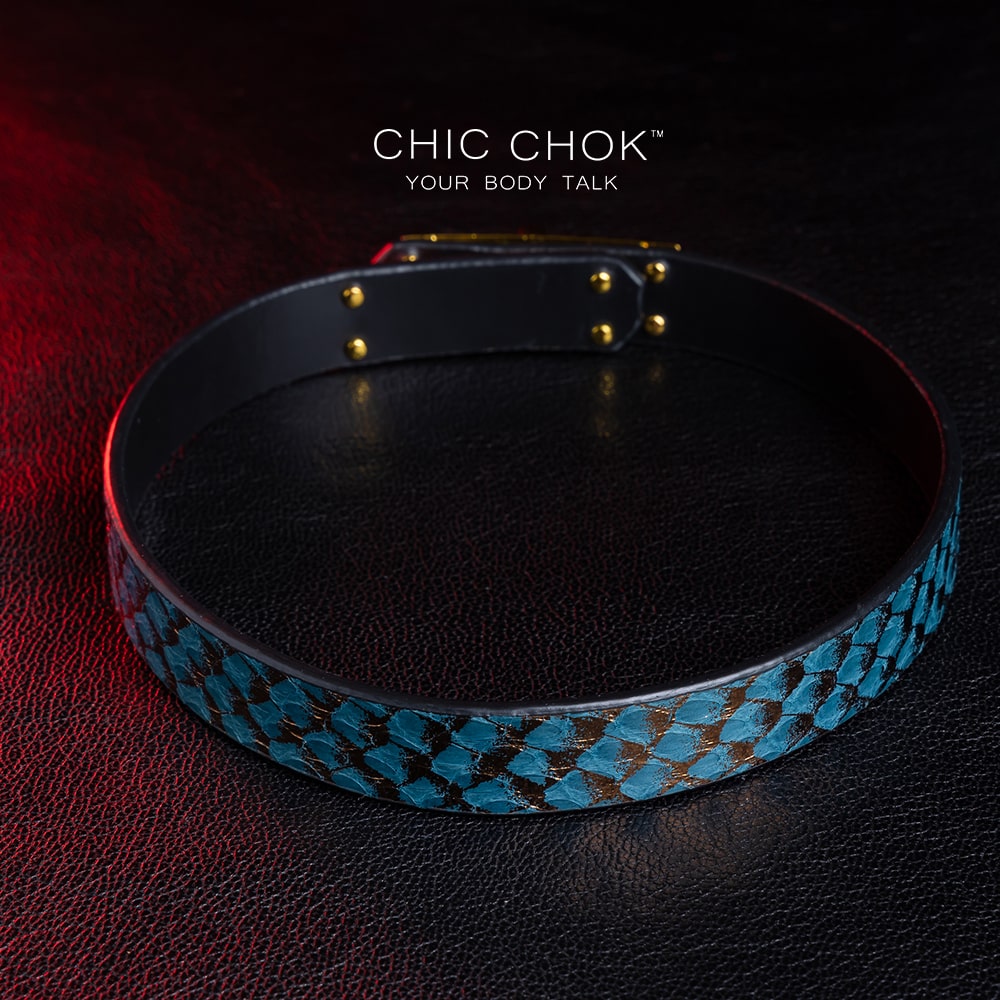 chicchok [Sapphire Moonlight] choker featuring a teal snakeskin-textured band with a gold-plated buckle clasp, displayed on a dark leather surface with dramatic red lighting accents, showcasing its luxurious and edgy design.