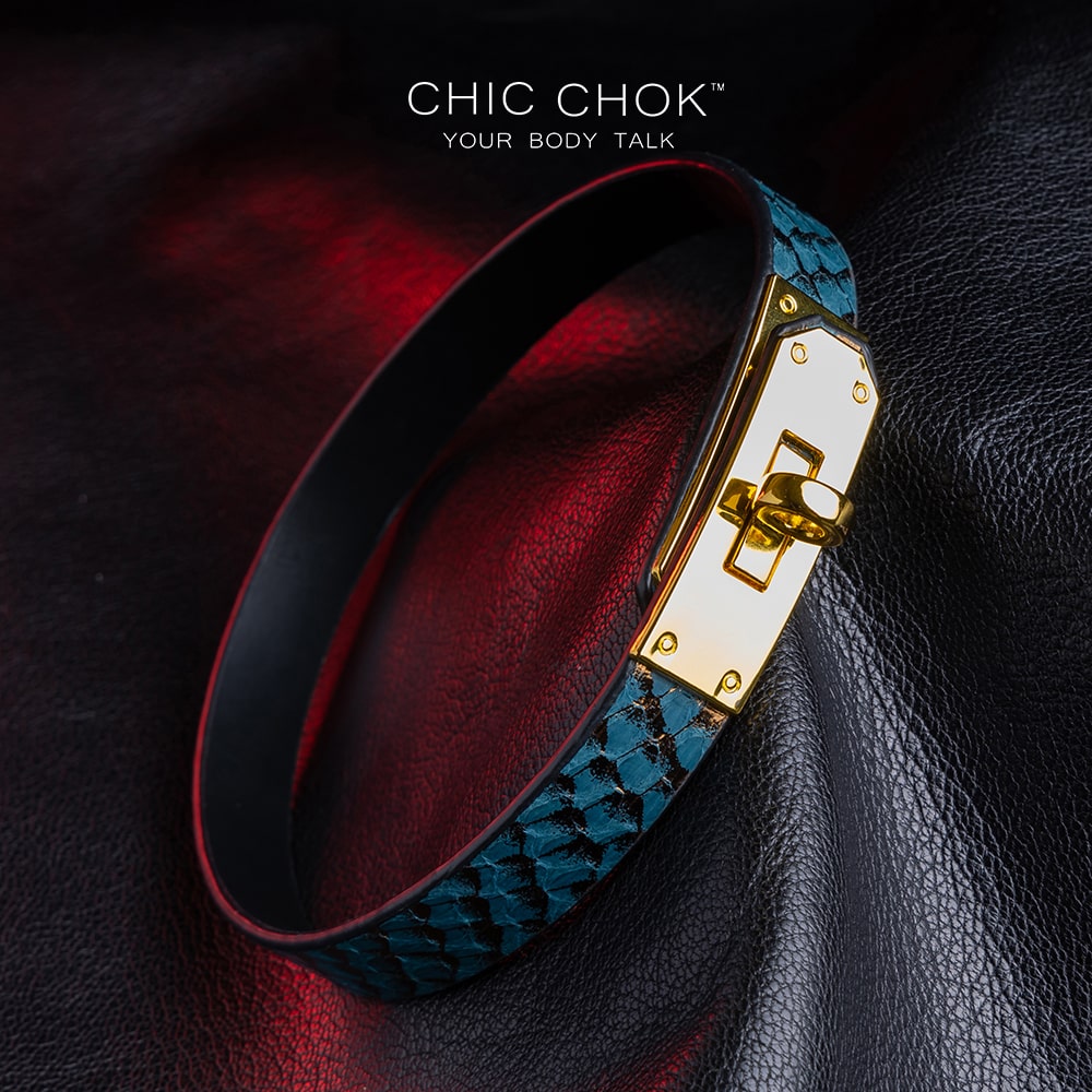 chicchok [Sapphire Moonlight] choker featuring a teal snakeskin-textured band with a gold-plated buckle clasp, displayed on a dark leather surface with dramatic red lighting accents, showcasing its luxurious and edgy design.