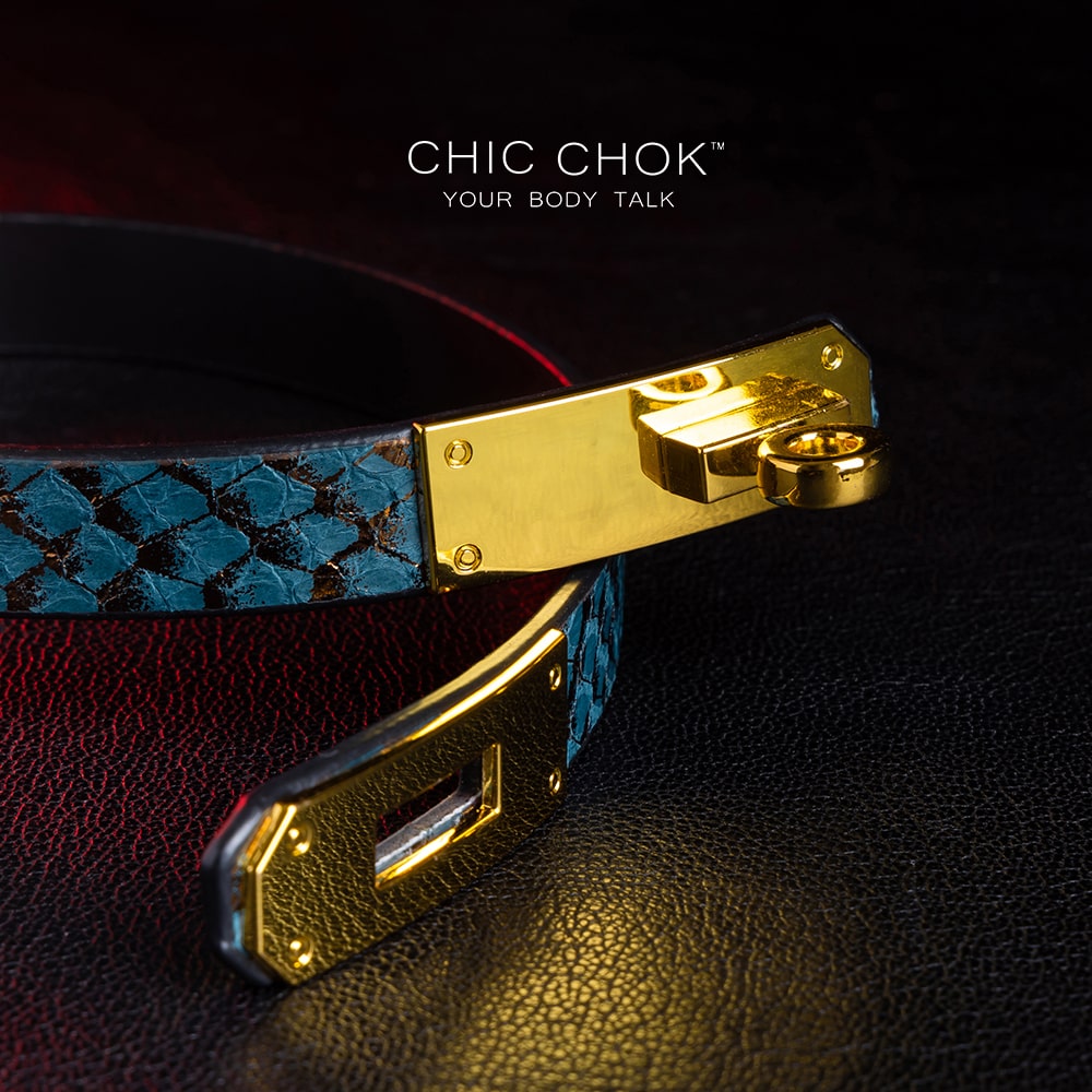 chicchok [Sapphire Moonlight] choker featuring a teal snakeskin-textured band with a gold-plated buckle clasp, displayed on a dark leather surface with dramatic red lighting accents, showcasing its luxurious and edgy design.