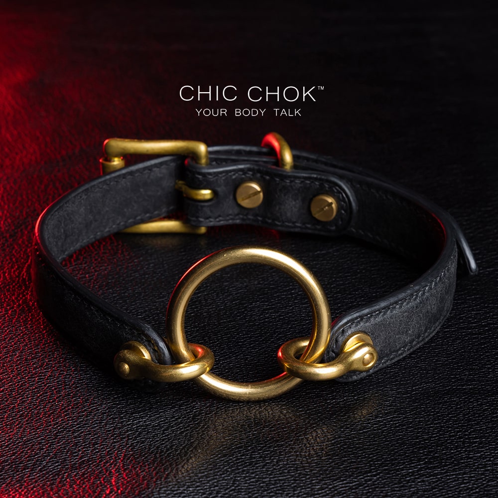 chicchok [Sinister] collar black leather choker featuring a central gold ring, gold-plated hardware, and adjustable buckle design, photographed on a textured black surface with warm lighting highlights