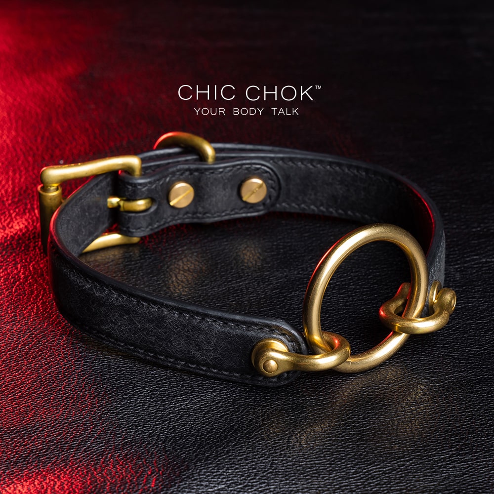 chicchok [Sinister] collar black leather choker featuring a central gold ring, gold-plated hardware, and adjustable buckle design, photographed on a textured black surface with warm lighting highlights