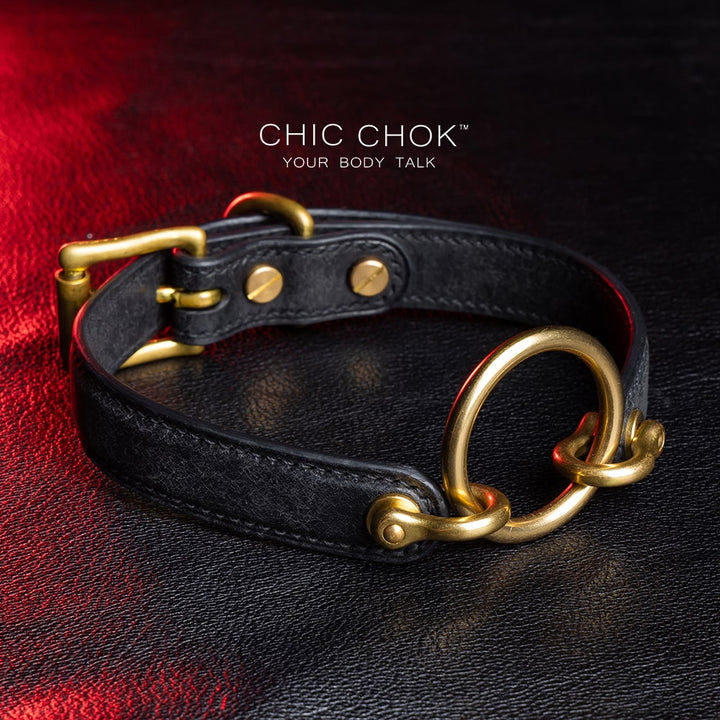 chicchok [Sinister] collar black leather choker featuring a central gold ring, gold-plated hardware, and adjustable buckle design, photographed on a textured black surface with warm lighting highlights