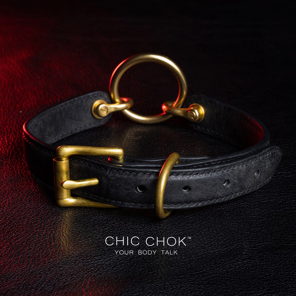 chicchok [Sinister] collar black leather choker featuring a central gold ring, gold-plated hardware, and adjustable buckle design, photographed on a textured black surface with warm lighting highlights