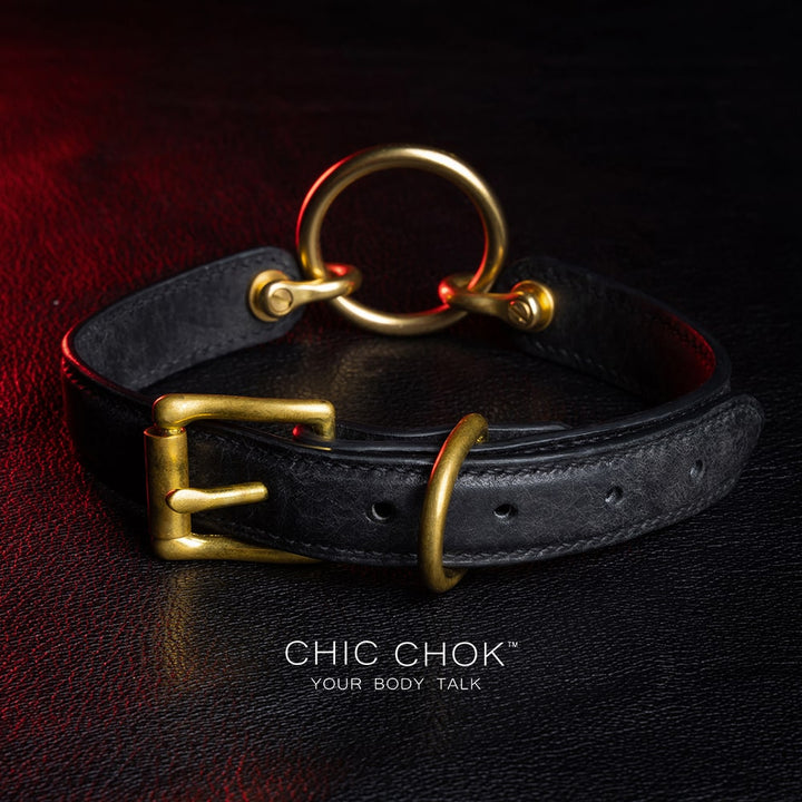 chicchok [Sinister] collar black leather choker featuring a central gold ring, gold-plated hardware, and adjustable buckle design, photographed on a textured black surface with warm lighting highlights