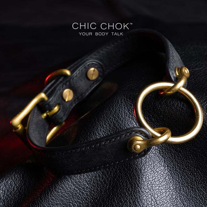 chicchok [Sinister] collar black leather choker featuring a central gold ring, gold-plated hardware, and adjustable buckle design, photographed on a textured black surface with warm lighting highlights