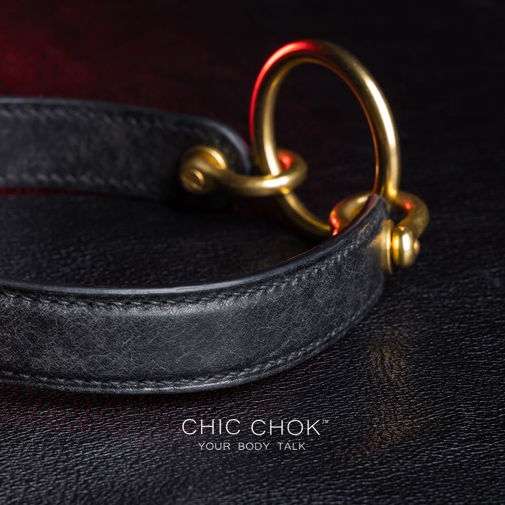 chicchok [Sinister] collar black leather choker featuring a central gold ring, gold-plated hardware, and adjustable buckle design, photographed on a textured black surface with warm lighting highlights